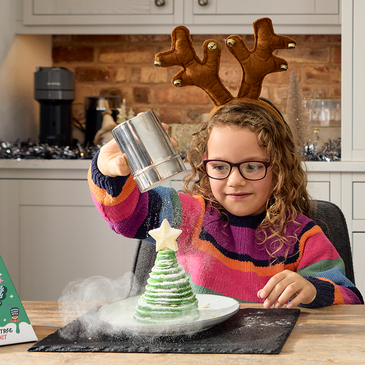 A child is pouring icing sugar on a tree-shaped pancake for a snowy finish, with the final product being a Christmas tree-shaped pancake on a plate.